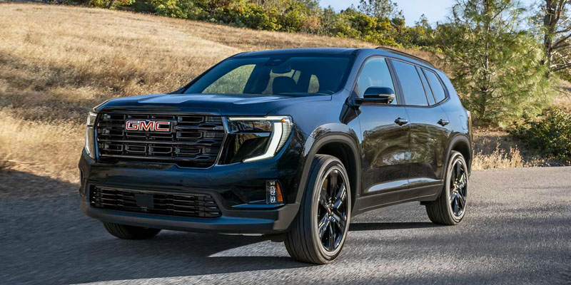 A black GMC Acadia SUV is being driven on a scenic road beside a newly-mowed golden field in a sunny countryside setting.