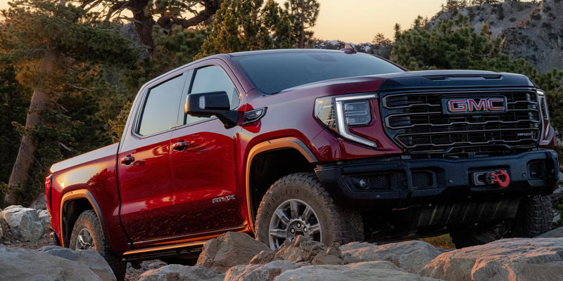 Red GMC Sierra AT4 pickup truck driving over large boulder terrain in a mountain landscape..