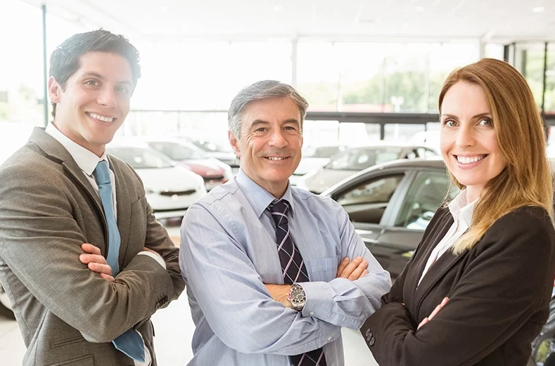 2 gentlemen and a woman sales persons are smiling at us from inside a sunlit GMC showroom with several vehicles on the floor behind them.