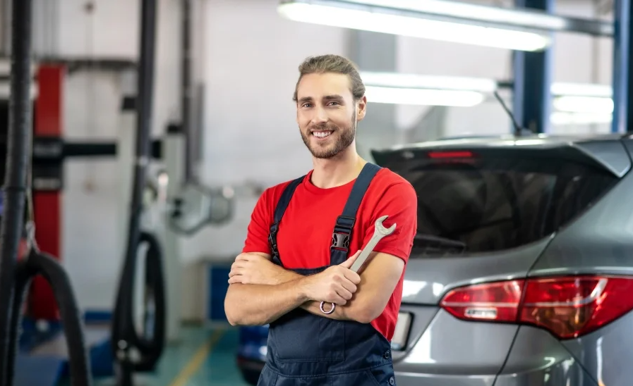 A GMC-certified service technician in a red t-shirt & overalls smiles at us while holding his wrench with crossed arms during an inspection of a vehicle in an automotive service center.